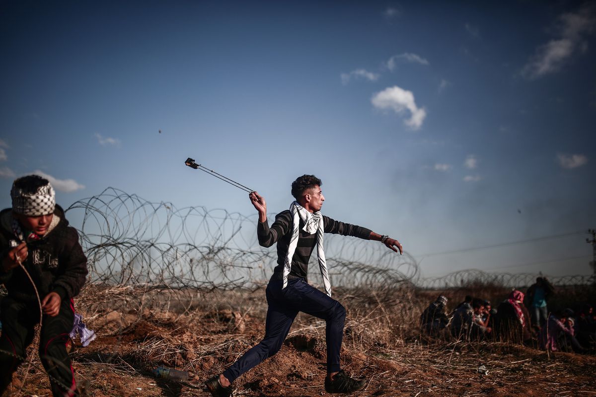 Under a blue sky, in a field surrounded by razor wire, a young man with a keffiyeh around his neck twirls a slingshot, aiming at troops off camera. His compatriots duck in anticipation of his release.
