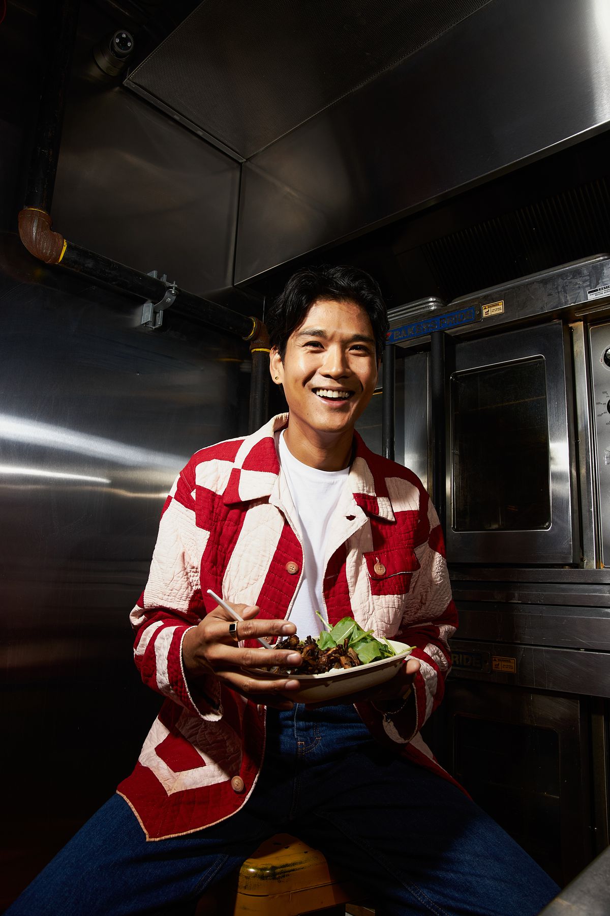 Vlady Rice, a Filipino chef known for his Waldy Cousin pop-up in New York City, stands in an industrial kitchen dressed in red and white.