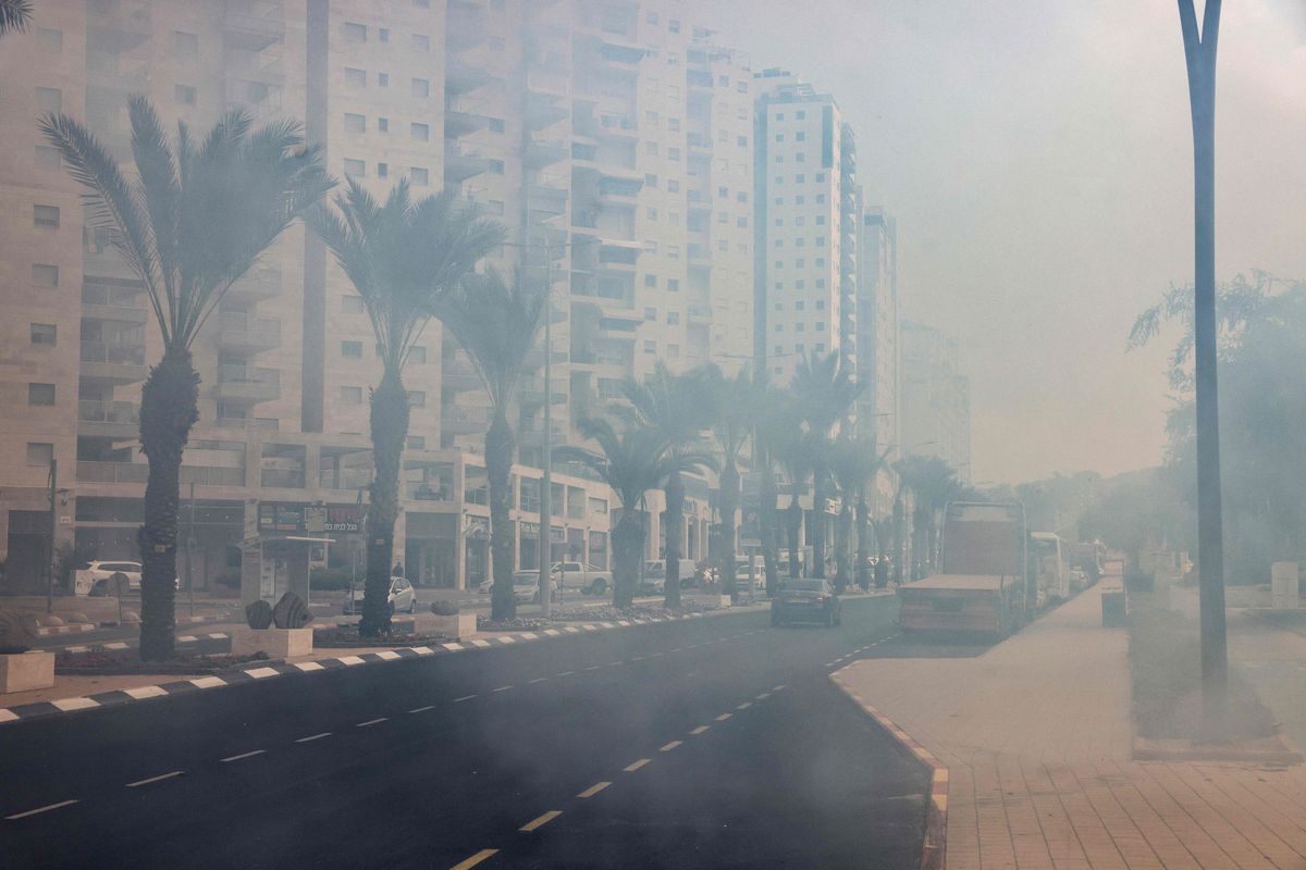 A photograph of a palm-tree-lined street in Ashkelon, in southern Israel. The light is dimmed by smoke from a nearby fire.