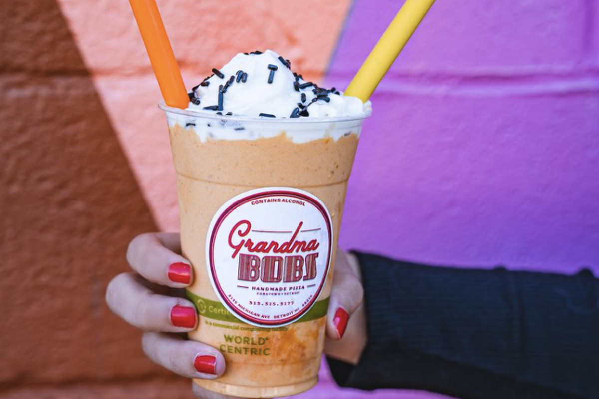 A hand holds the pumpkin vegan shake in a plastic cup topped with whip and sprinkles. It has two plastic straws sticking out from it.