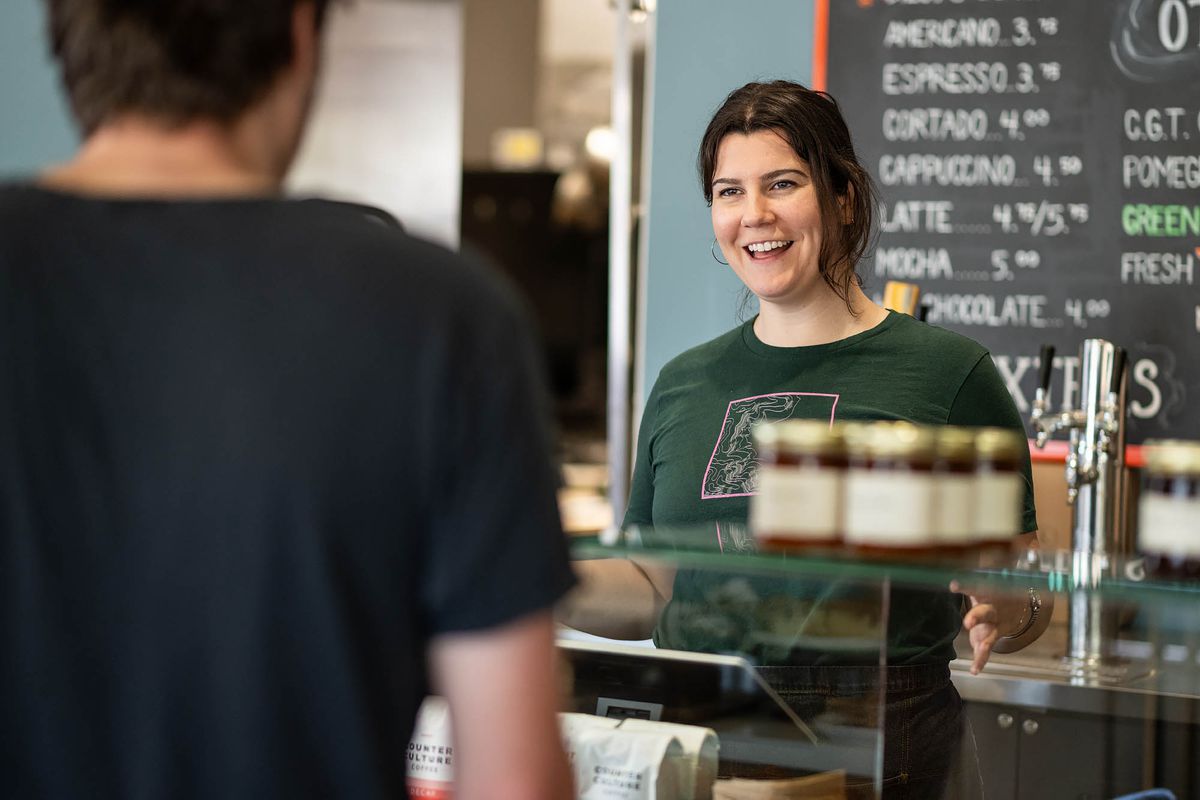 A dark-haired woman smiles at a customer, back to camera, from inside a sunny morning bakery in Los Angeles.