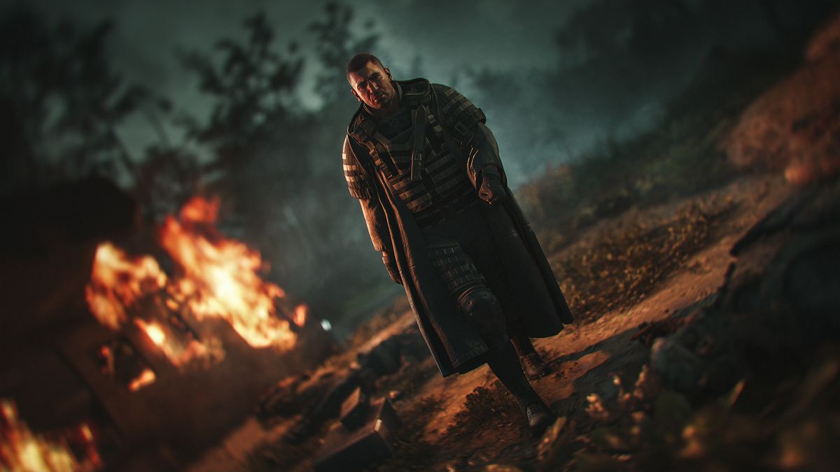 The villain Cole Walker cuts a menacing figure on a battlefield with fires glowing in the background