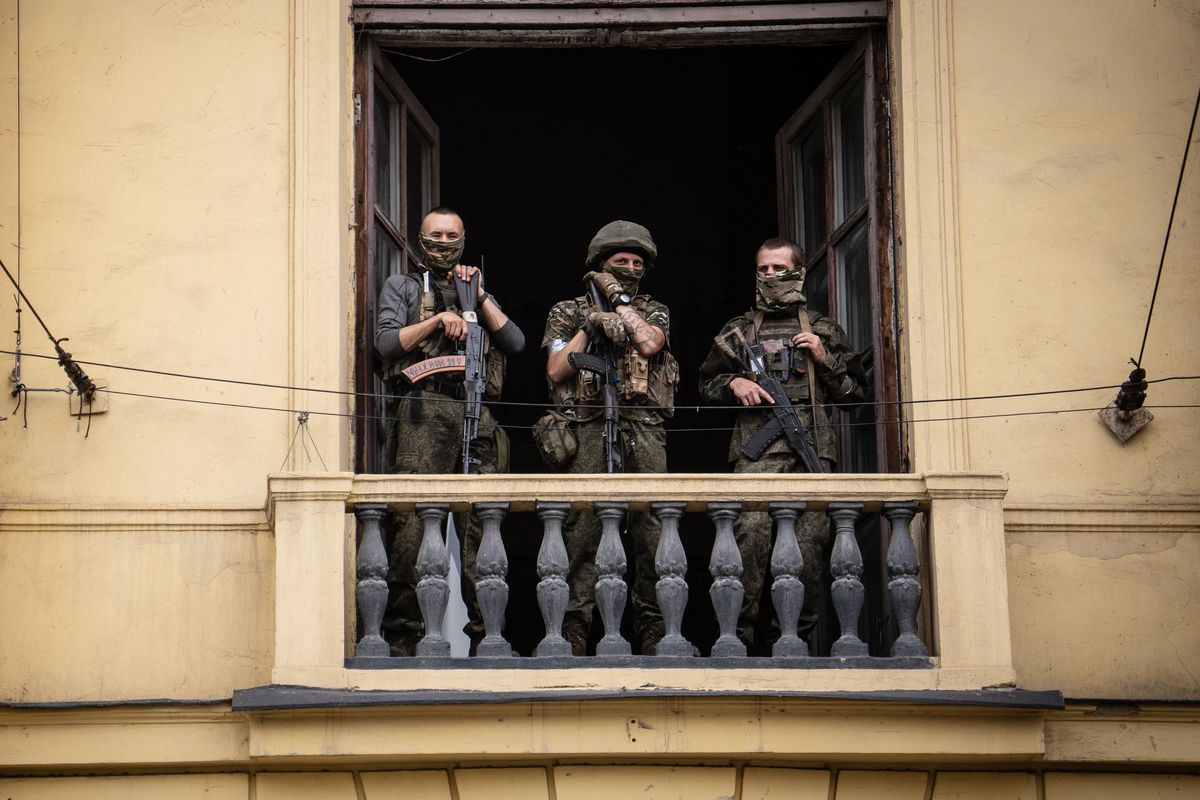 Three heavily armed men in camouflage military fatigues and army-green face coverings stand on a balcony.