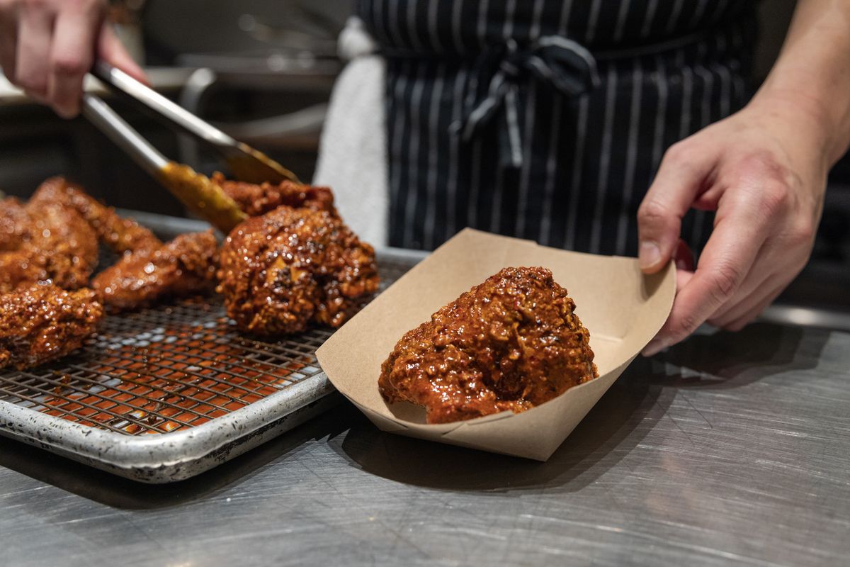 A chef is shown dunking fried chicken in a brown chile sauce and placing the poultry in a cardboard serving vessel