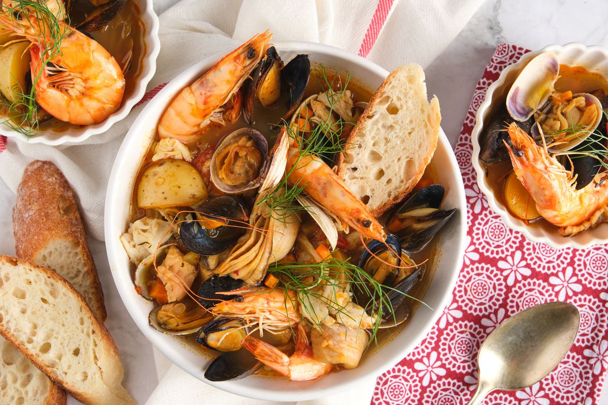 A big bowl of bouillabaisse sits on a table with slices of baguette and smaller bowls of shrimp.