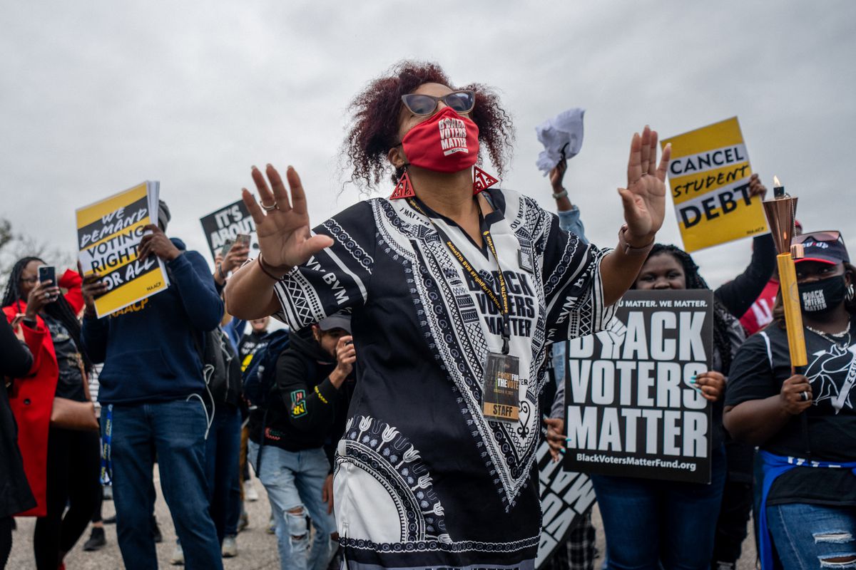 A black women in a red mask reading “Black Voters Matter” and wearing a black and white dashiki with the same message printed across the chest, raises her arms as she leads a group of Black demonstrators, many holding signs reading “Black Voters Matter” and “Cancel Student Debt.”