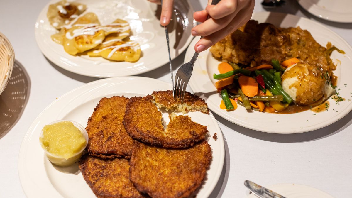 A pair of white hands use a fork and knife to slice off a piece of potato pancake on a plate with apple sauce. It’s surrounded by plates of pierogi and city chicken with mashed potatoes and steamed veggies.