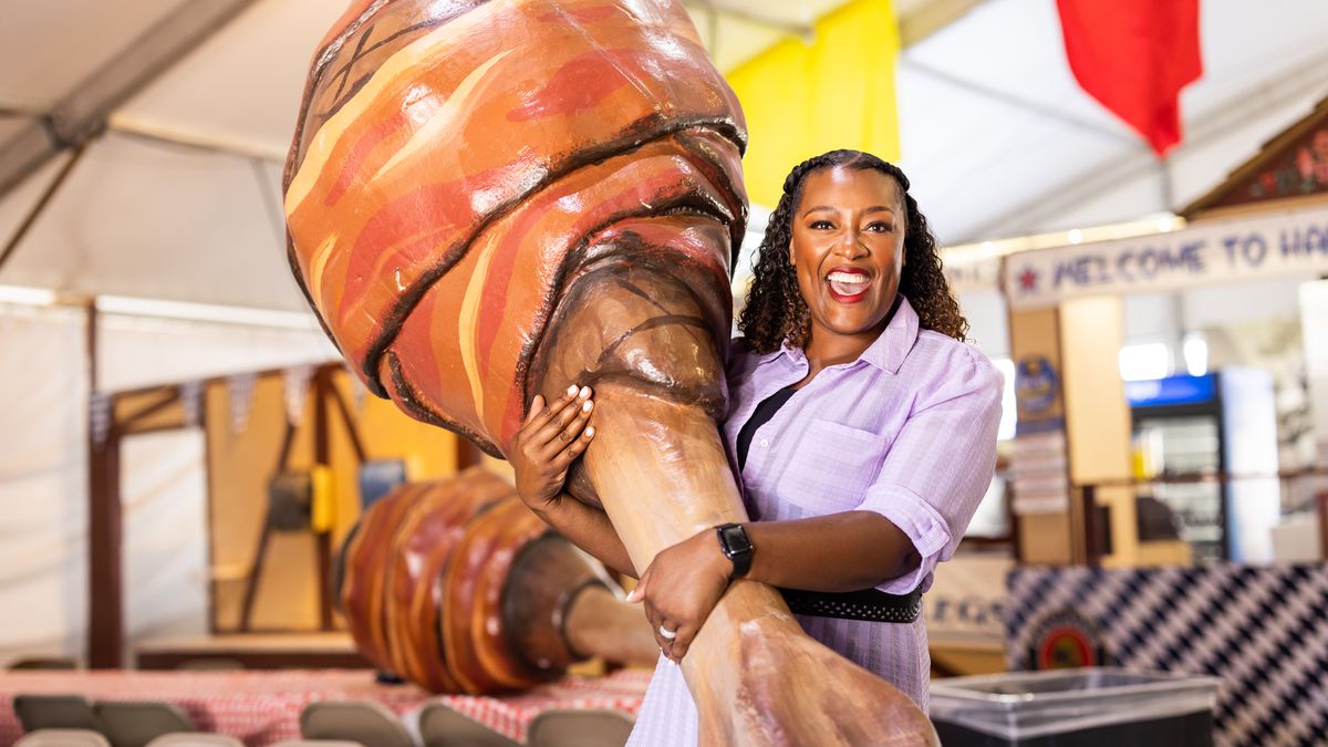A smiling woman holds an oversized sculpture of a turkey leg wrapped in bacon.