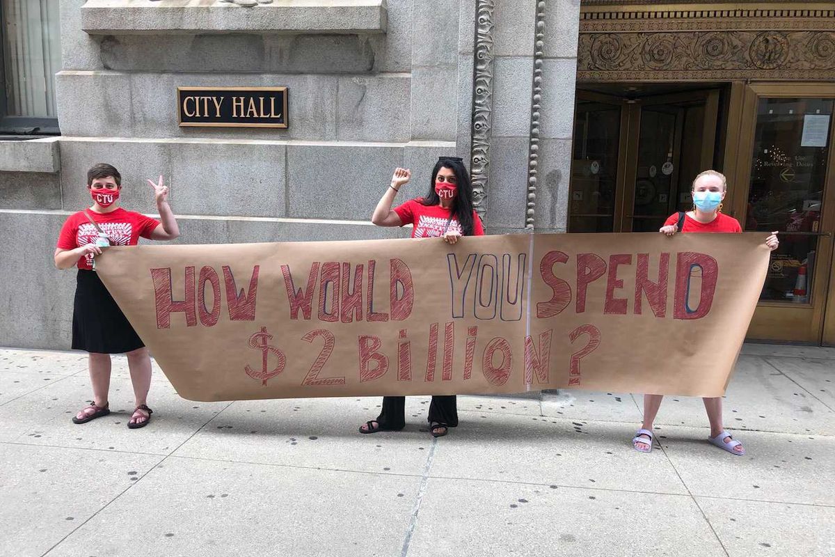 Three members of the Chicago Teachers Union stand outside City Hall. They wear red t-shirts and hold a brown sign that says, “How would YOU spend $2 billion?”