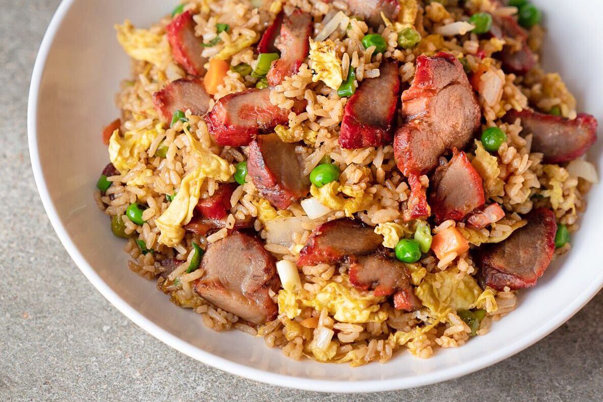 A shallow white bowl filled with fried rice, green peas, and chunks of barbecue pork, along with other vegetables, that is placed on a light gray table