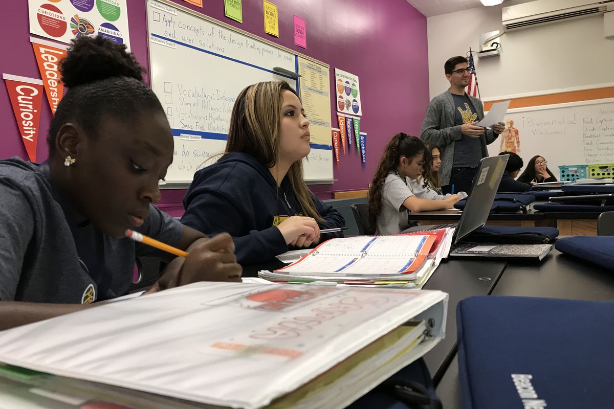 Six students at Kepner Beacon Middle School sit at long tables and work on an assignment. One girl holds a pencil. A male teacher stands in the background. Behind them is a purple wall with banners saying ’Curiosity” and “Leadership,” a whiteboard, and a poster listing character traits.
