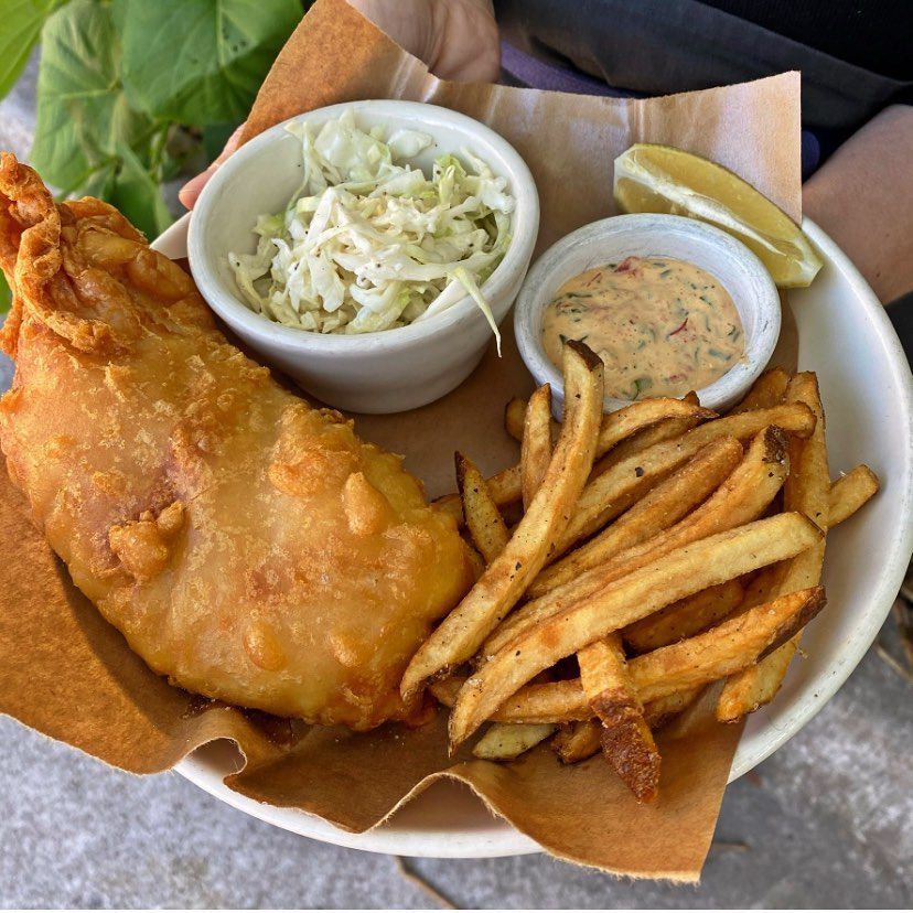 A plate with brown paper of fried fish, long fries, and saucers of coleslaw and an orange sauce.