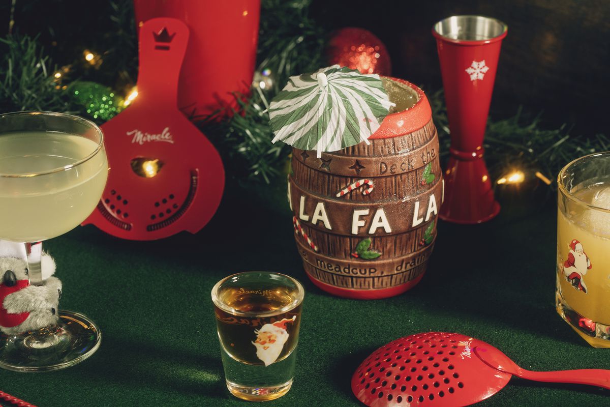 A holiday-themed tableau includes a mug shaped like a barrel that says “Fa-La-La” with a cocktail umbrella, a Santa shot glass, and red cocktail mixing tools.