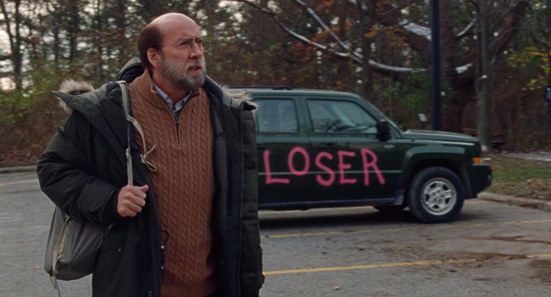 A bearded, bald, middle-aged man stands in a parking lot, looking confused. Behind him is a car with “LOSER” spray-painted onto it.