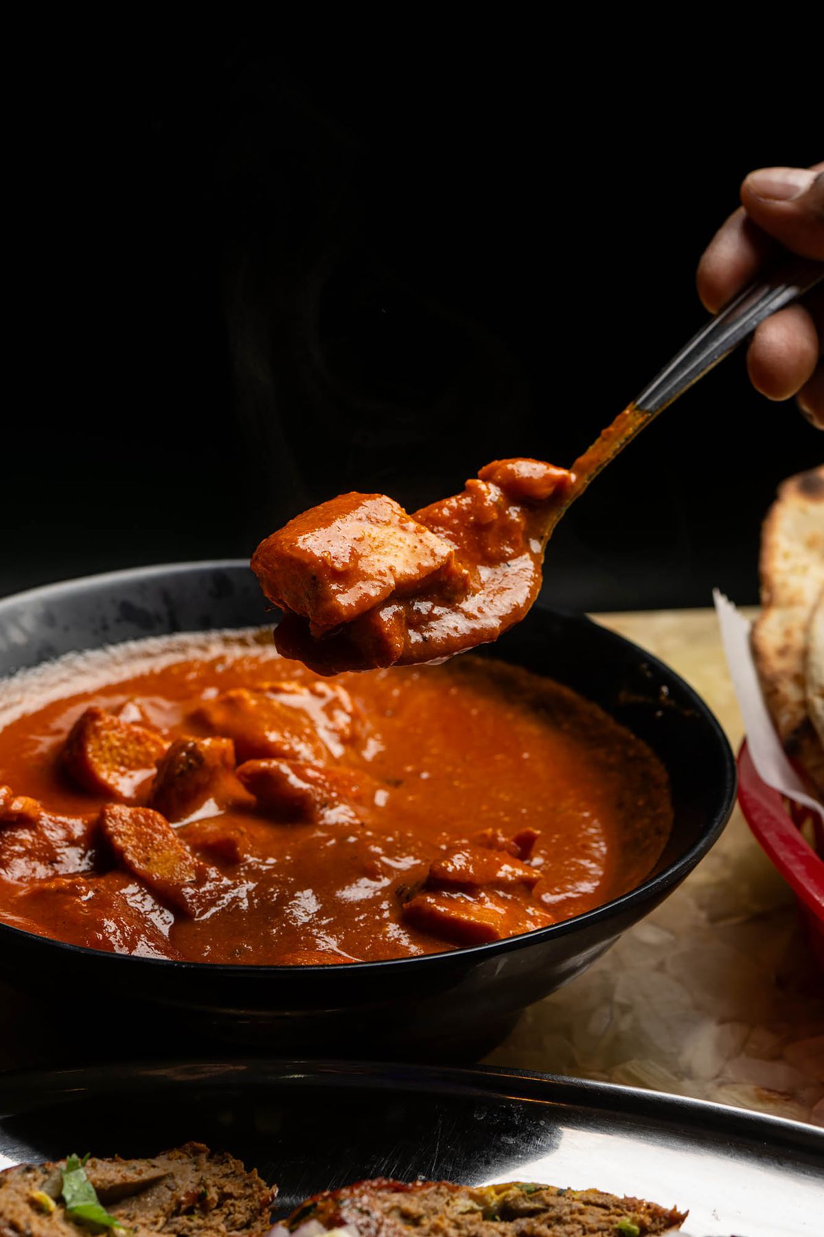 A black bowl of Chicken tikka masala with a close up of the dish on a spoon. 