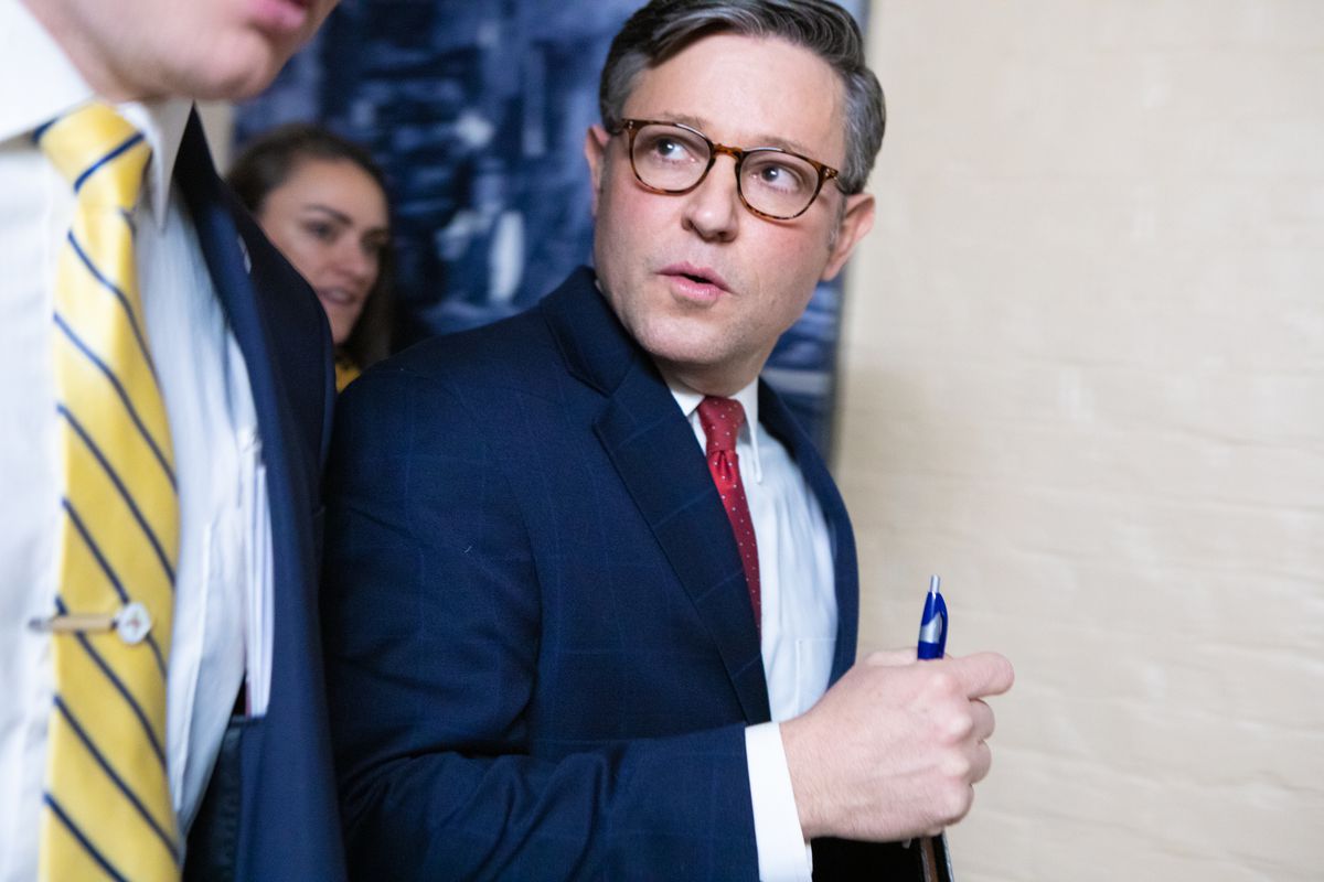 Mike Johnson pictured wearing a navy suit jacket, red tie, and glasses. He appears to be holding a blue pen in his hand.