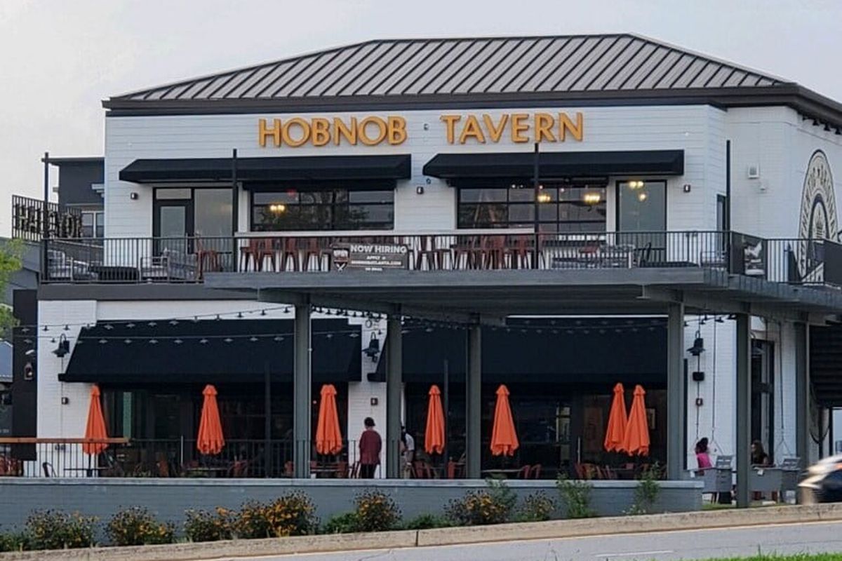 The two-story Alpharetta location of Hob Nob Neighborhood Tavern, a white building with black trim and roof, two patios, and orange umbrellas on the ground floor patio 