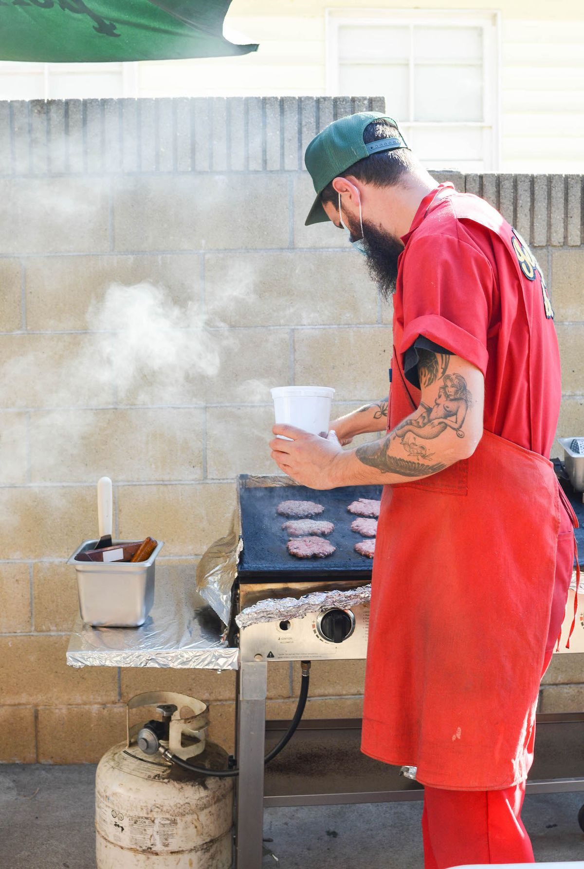 A man in a red jumpsuit smashes burgers on a smoky griddle.