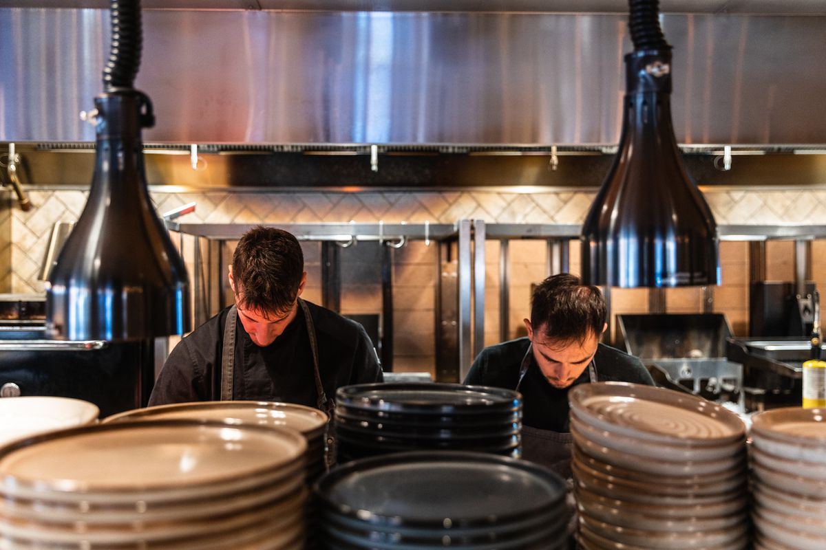 Two chefs at work under heat lamps behind stacks of plates.