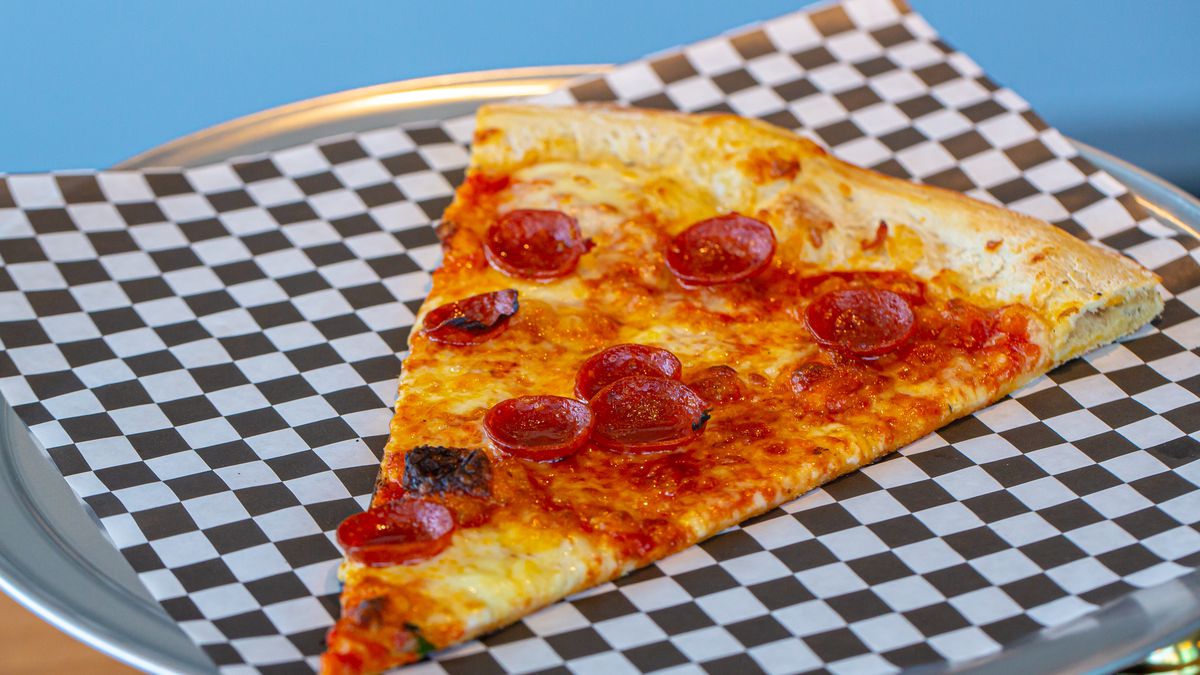 A slice of pepperoni pizza atop checkerboard tissue paper on a round pizza tin