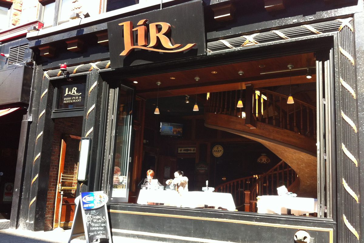 Exterior of a bar with signage reading “Lir.” The exterior is dark wood, and a giant nearly floor-to-ceiling window takes up most of the space, open to the sidewalk.