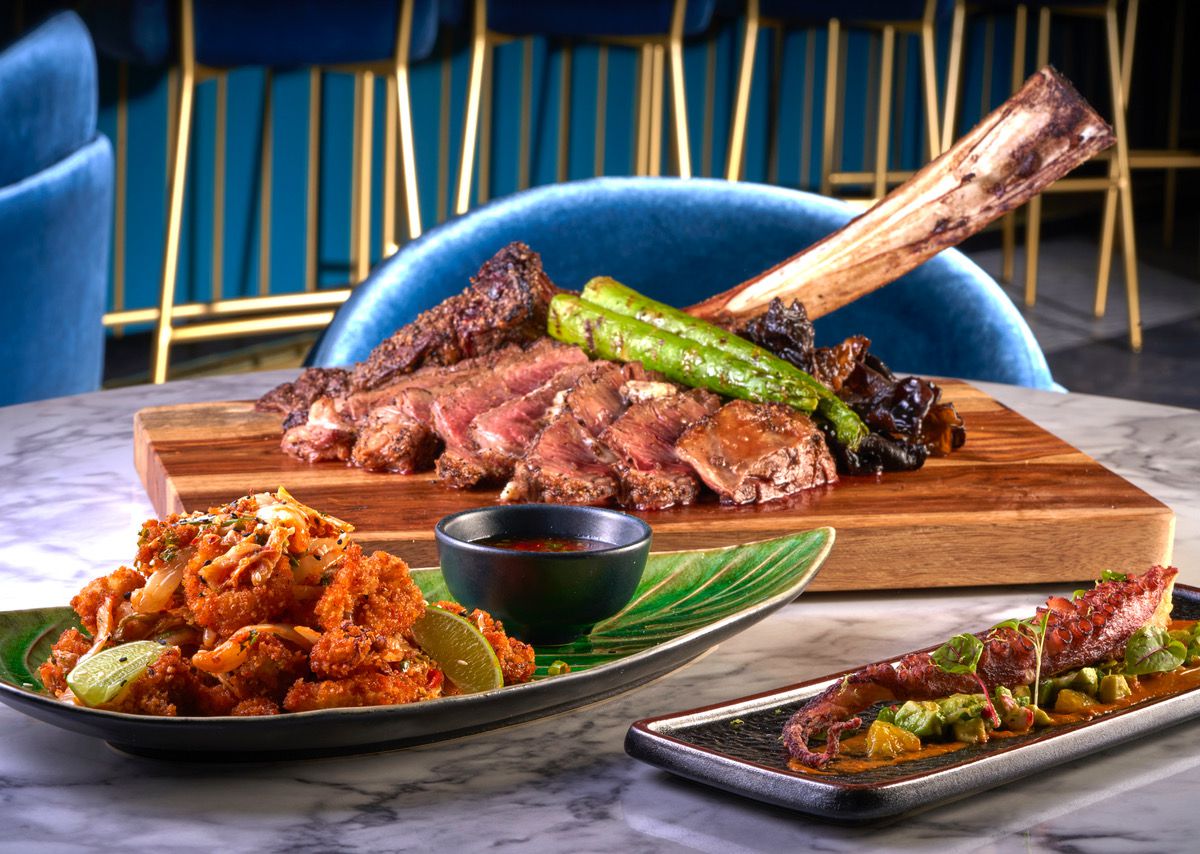 On a table are a wooden slab with a bone-in steak, plus two plates of appetizers. Blue velvet furniture is visible in the background.