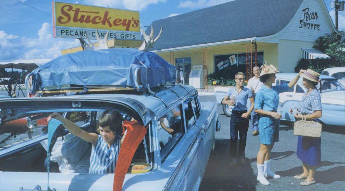 A 1970s advertisement for Stuckey’s truck stop chain with people standing in the parking lot chatting and a little girl hanging out of the back of a parked blue station wagon packed for a road trip. 