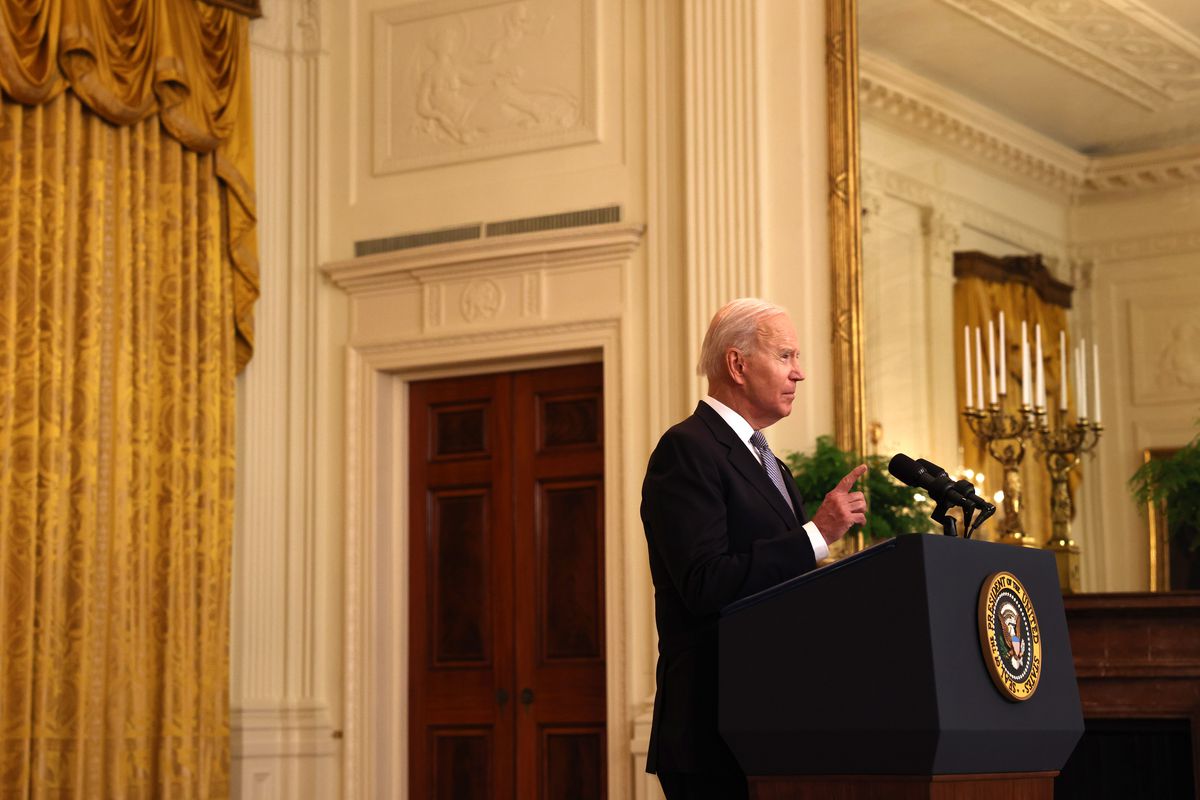 President Joe Biden speaks at a podium marked with the Presidential seal in a room with golden drapes and a large candelabra behind him.
