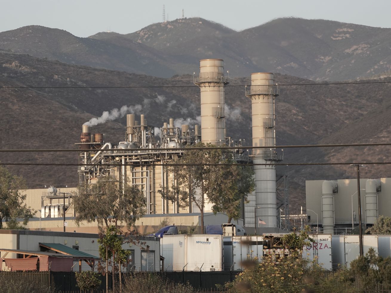Amid rolling, verdant hills clothed in smog, sit two wide smoke stacks, positioned just to the right of thin metal towers and thinner smokestacks spewing fumes. A tangle of black wires run horizontally across the photo’s foreground.
