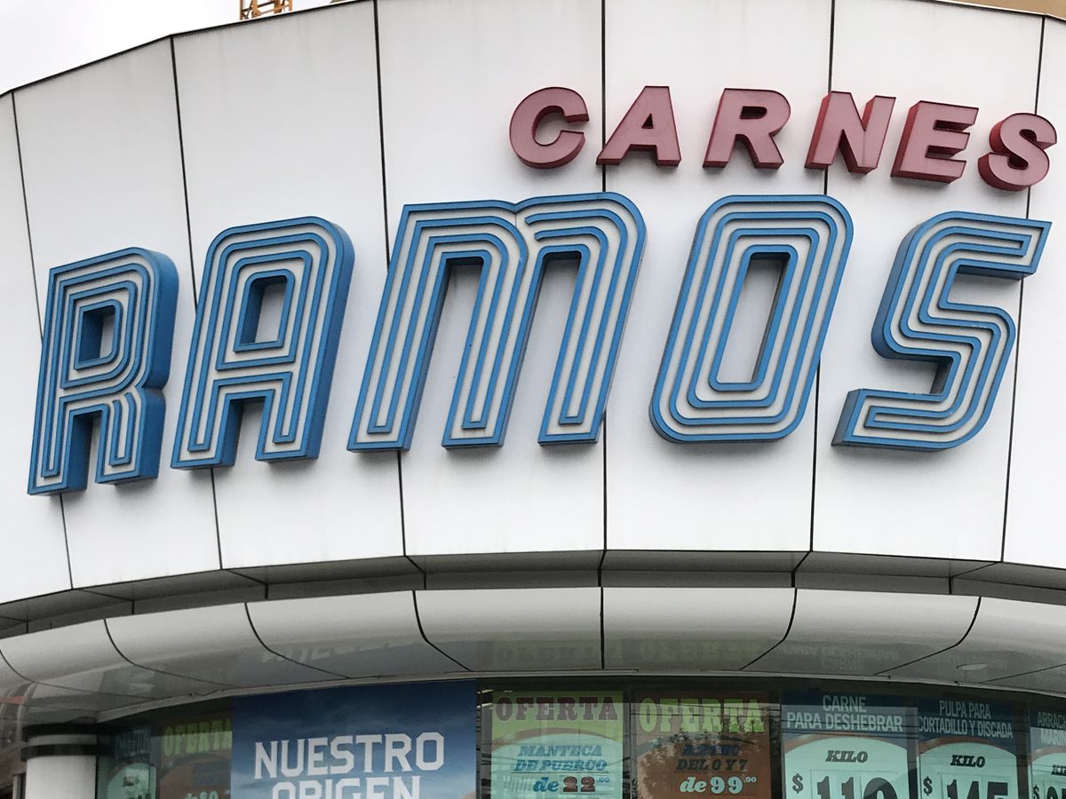 The exterior of the Carnes Ramos butcher shop with large letters built into the second floor facade above windows plastered with posters advertising meat sales
