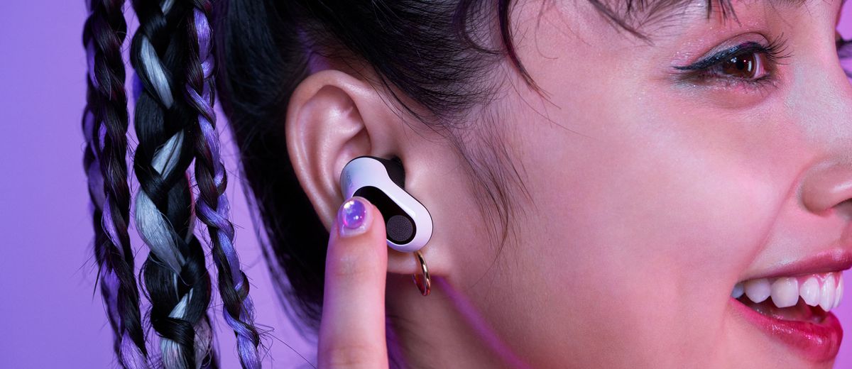 A photo taken by Sony showing a smiling person wearing its Inzone Buds wireless earbuds. They are holding a finger closer to their ear to adjust a setting on the earbuds.