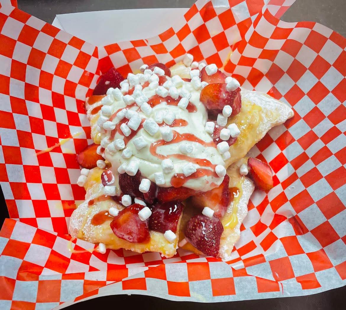 A trio of sopapillas sit in a cardboard boat on top of red-checked paper. They’re covered with strawberries, creme, and tiny dehydrated marshmallows.