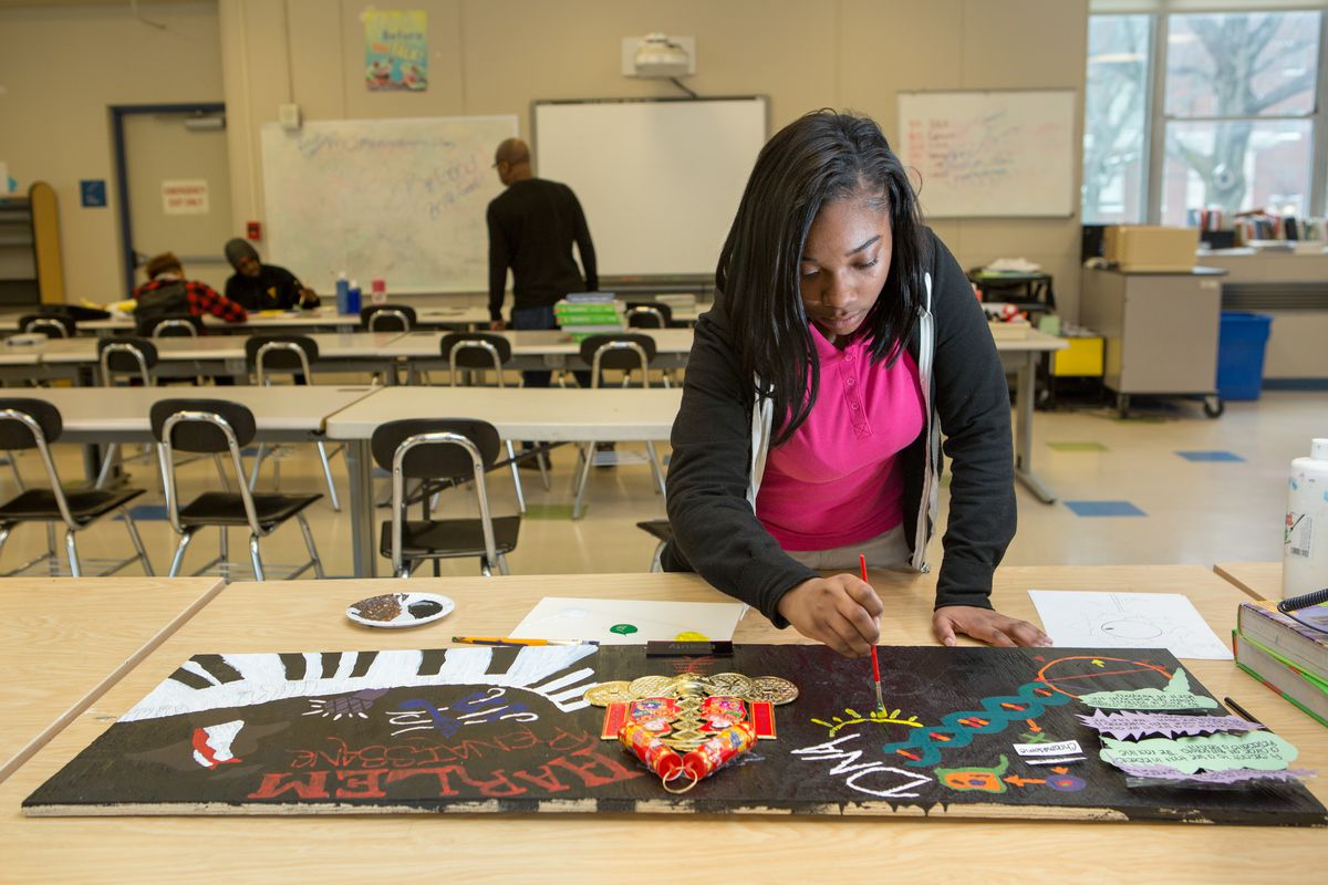 A girl wearing a black hoodie and a pink polo shirt works on an art project while other students speak with a teacher on the other side of the classroom.