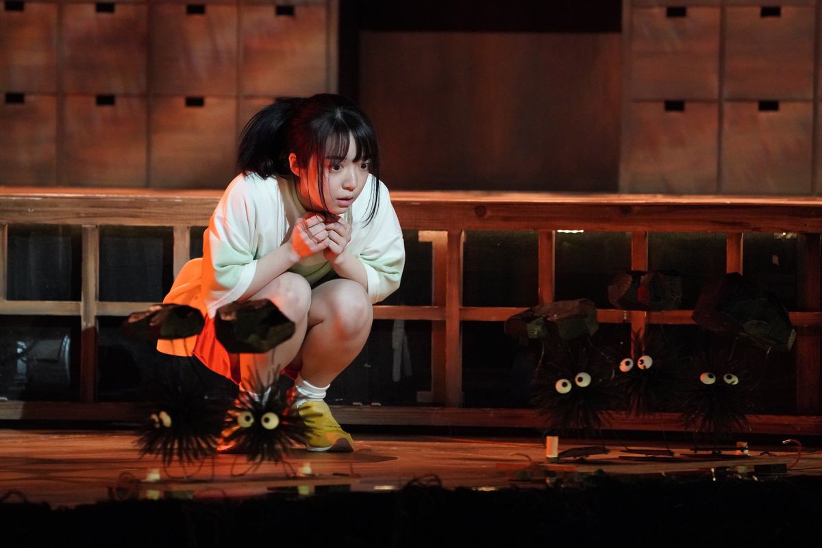 Chihiro crouches and looks at the puppets of coal-carrying soot sprites moving across the stage in Spirited Away: Live on Stage