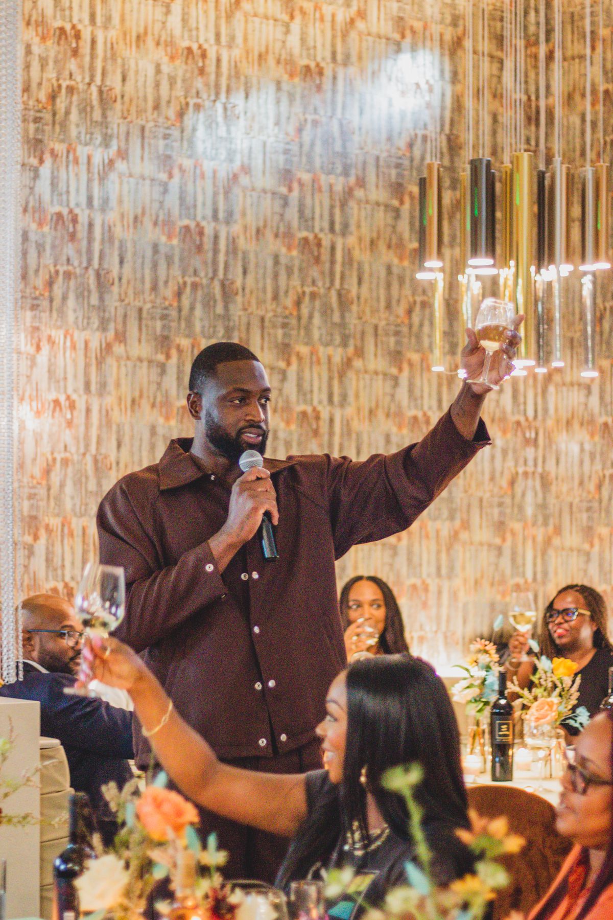 Man holds up a wine glass in a toast in one hand and speaks into a microphone.