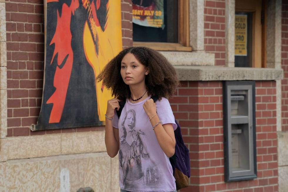 Sarah (Nico Parker) walking. She has a purple Halican Drops shirt on, with a woman holding a microphone and singing