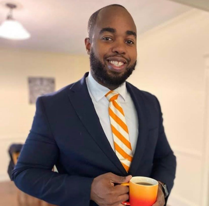 Teacher Cartavius Black holds a cup of coffee standing in a living room wearing a suit and tie.