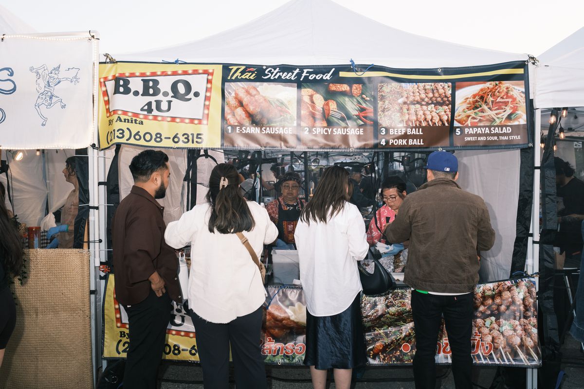 A group of people wait in front of a food stand that has signs for barbecue items.