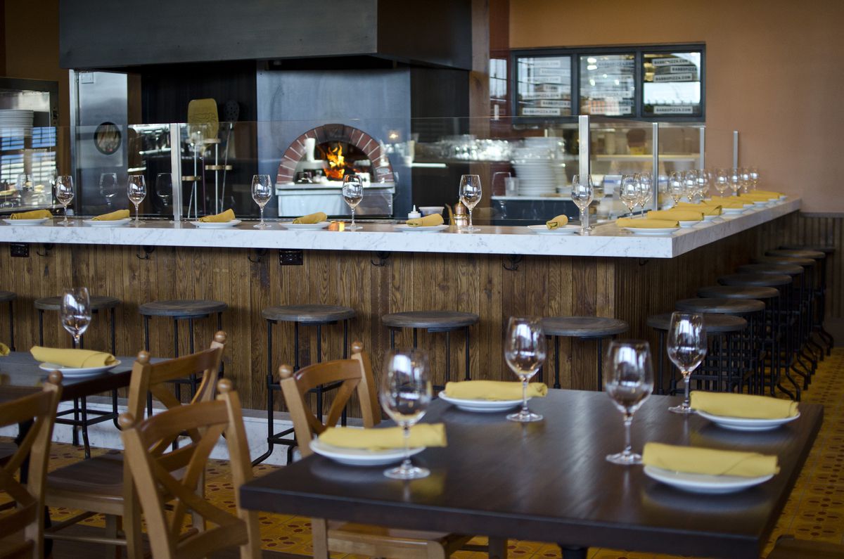 A pizza oven burns in the background of this photo of part of the Babbo Boston interior. Counter seating surrounds the oven, and there are regular dining tables in the foreground.
