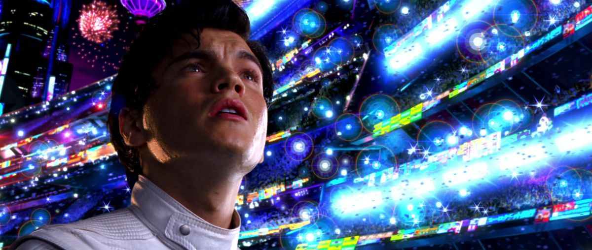 A young man (Emile Hirsch) with dark hair in a white collar racing jacket looks up at a stadium filled with blinding lights with fireworks in the distance.