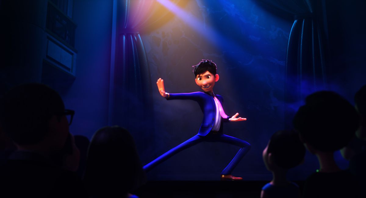 Din, in a blue suit and pink tie, poses in a dramatic kung-fu-style pose onstage in front of an audience in Wish Dragon