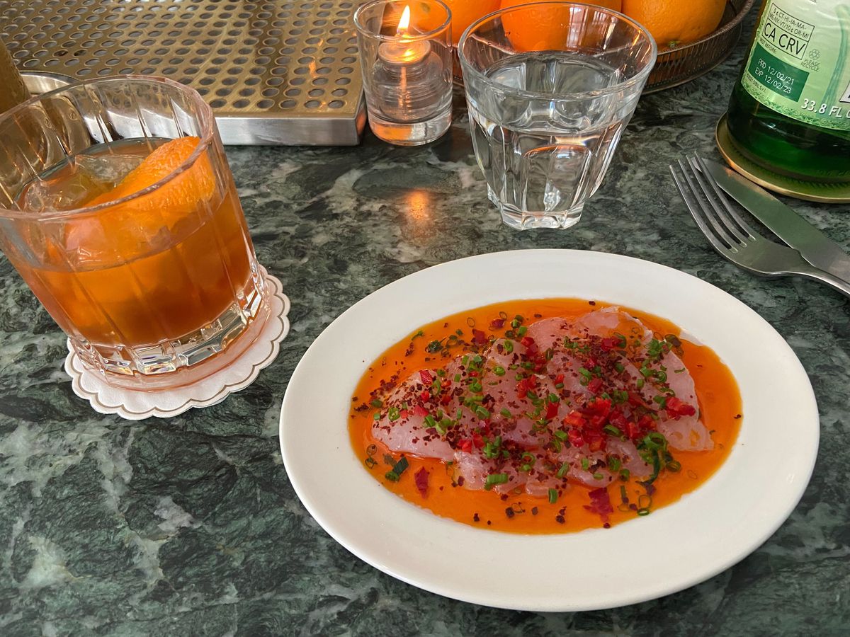 A green marble countertop features an orange cocktail and a white plate with a pink raw fish crudo sitting in a orange oil bath with green and red garnishes.