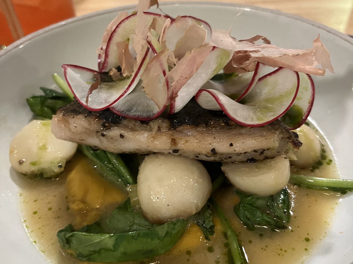 A piece of fish topped with thin-sliced radishes and sitting on a bed of vegetables and broth.