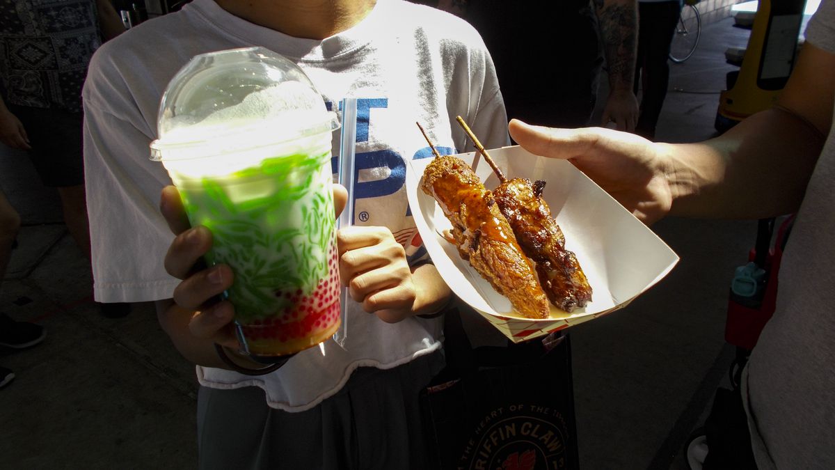 Someone holding a multicolored beverage in a plastic cup and a hand holding a paper tray of skewers.