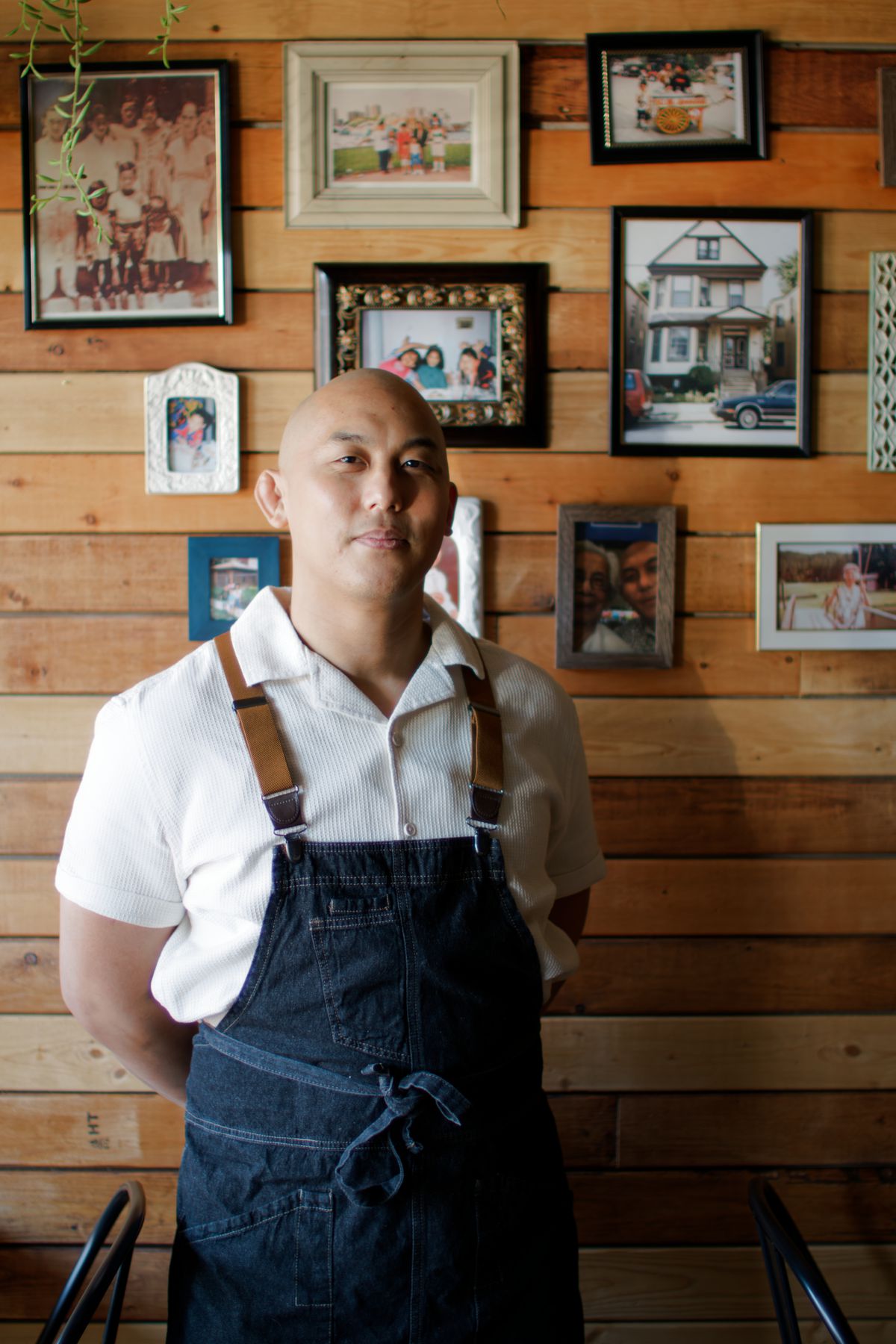 A male chef in a blue apron poses with a serious expression in front of a wood wall decorated with framed family photos. 