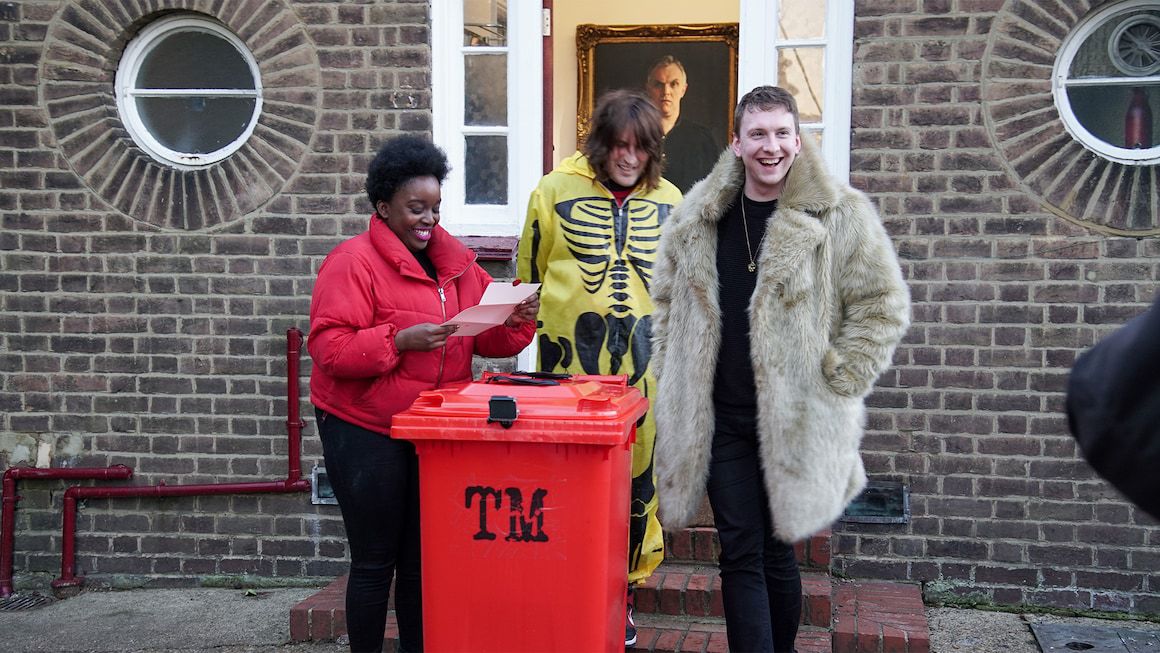 Lolly Adefope, Noel Fielding, and Jo Lycett smile together while reading a task above a red trash can outside the Taskmaster house. Adefope wears a bright red coat, Fielding wears a skeleton costume, and Lycett wears a big fur coat.