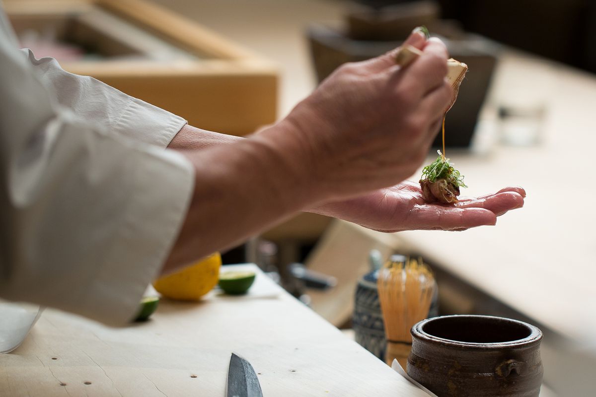 Chef Masa Takayama prepares sushi with his hands over a blond wood counter.