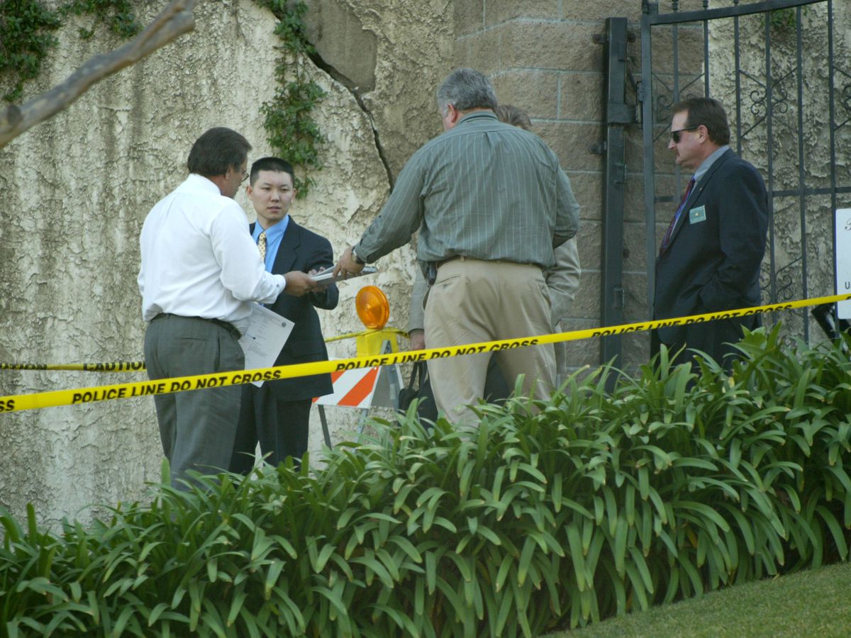 A group of men stand outside Phil Spector’s home which is the crime scene of Lana Clarkson. They are standing behind yellow crime scene tape which is stretched across a driveway.