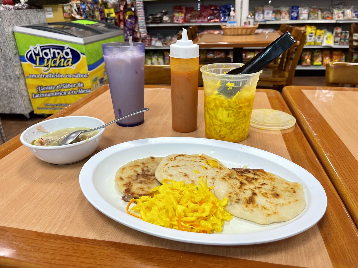 Three pupusas sit on a plate beside a clump of cabbage, a bowl of chicken soup, a glass of horchata, and a container of spicy sauce.