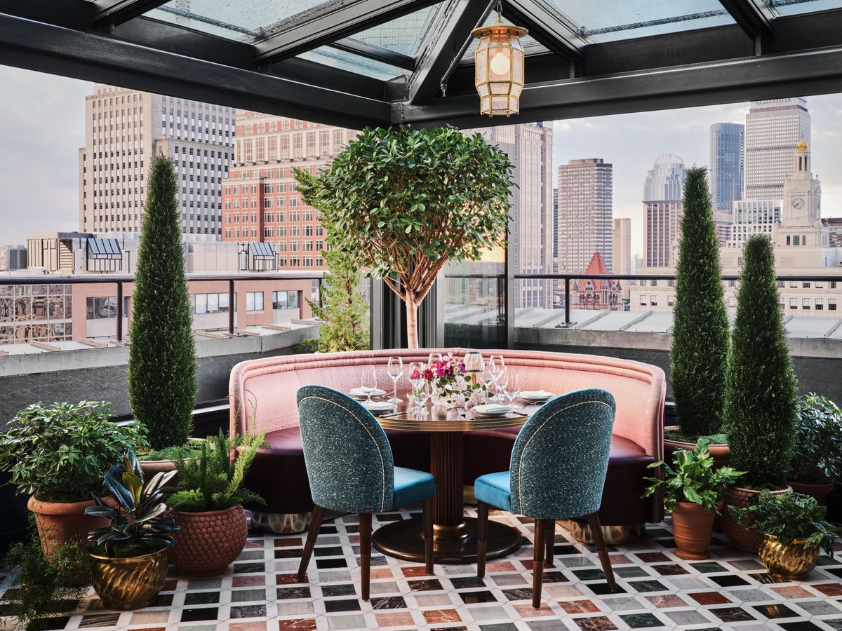 A round restaurant table features rose-colored booth seating and teal chairs. The floor is marble with a multi-colored diamond pattern, and the table is surrounded by plants. Views of Boston’s Back Bay are clearly visible behind the table.